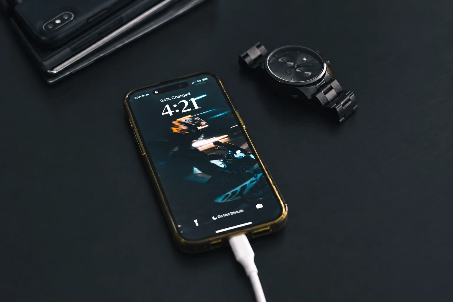 A phone connected to a charging cable beside a wristwatch on a dark table.