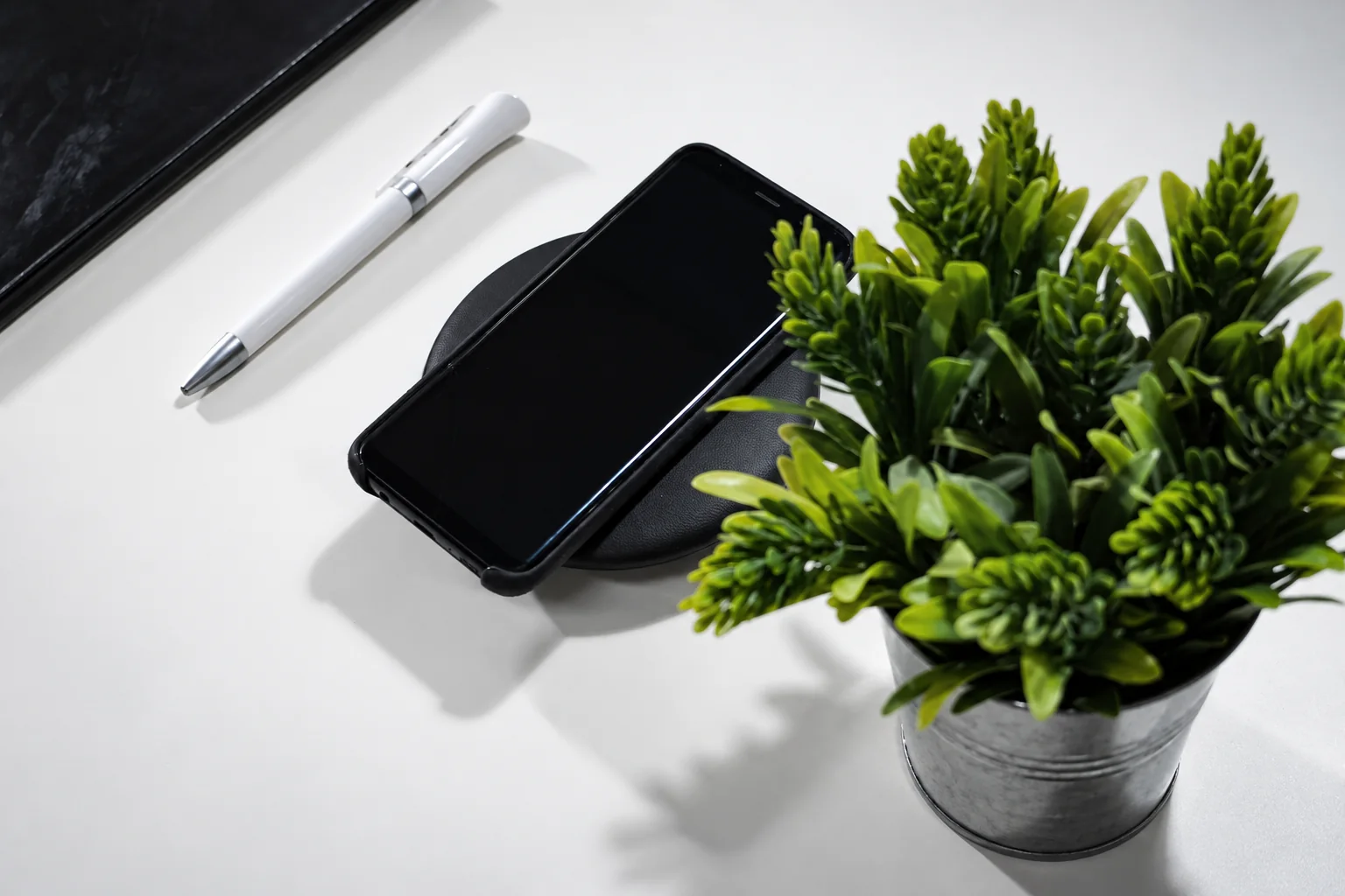 A phone on a round wireless charging pad beside a pen and plant.